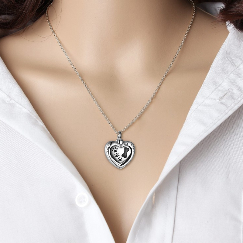 silver heart urn with black dog paw prints on it and the words "always in my heart" on the front  hanging on a silver chain hanging on a woman's neck.