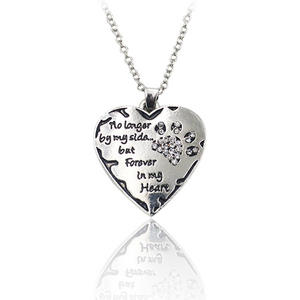 silver heart pendant with  a silver crystal paw print on its side in the top corner and the words"no longer by my side... but forever in my heart" written next to the paw print hanging on a silver chain.