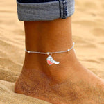 Sand filled image with a ladies foot. The foot has a beaded chain anklet on the ankle and a little red and white robin hanging on it.  The ladies leg has trousers on it. 